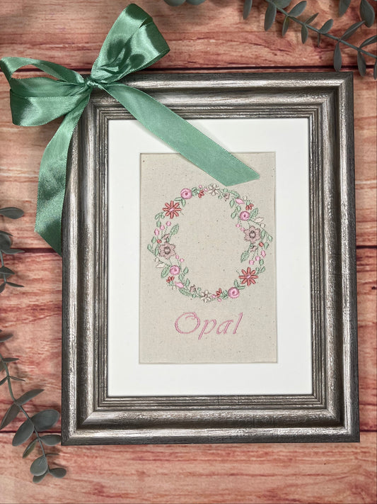 Framed artwork with floral wreath and 'Opal' text on a wooden surface with green ribbon and leaves.