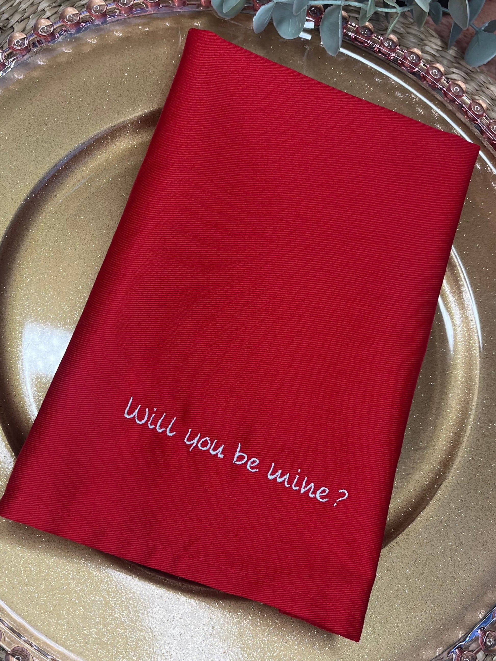 Red napkin with 'Will you be mine?' on a gold plate with silverware, surrounded by greenery.