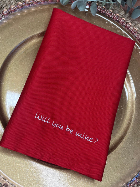Red napkin with 'Will you be mine?' on a gold plate with silverware, surrounded by greenery.